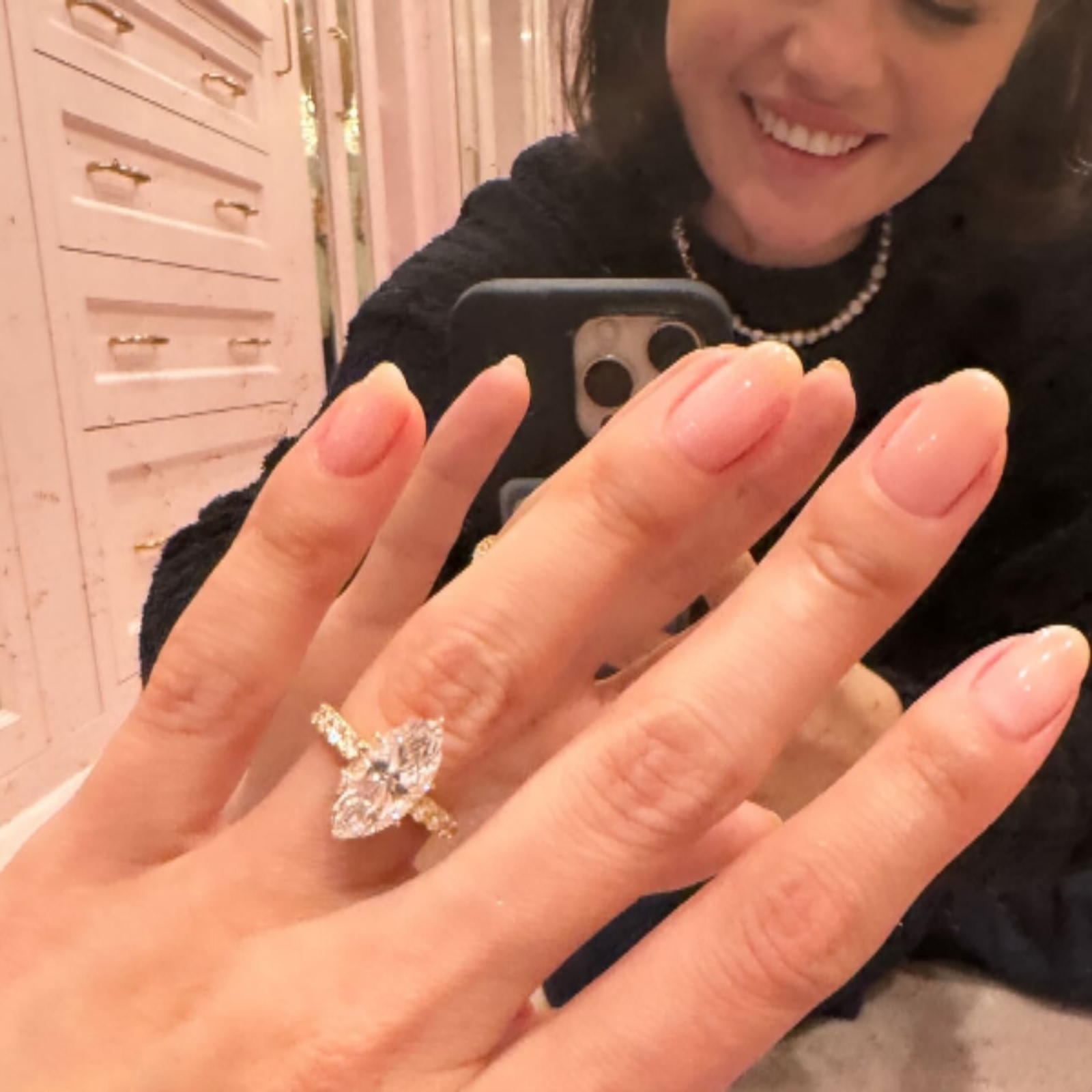 Strahlende Frau zeigt ihren eleganten Verlobungsring mit einem Diamanten im Marquise-Schliff und einem goldenen Pavé-Band. Dieses exquisite Design können Sie bei NÓIR.com erwerben – wahlweise mit natürlichen oder labgrown Diamanten, garantiert zum besten Preis. Perfekt für Paare, die einen modernen und nachhaltigen Verlobungsring suchen